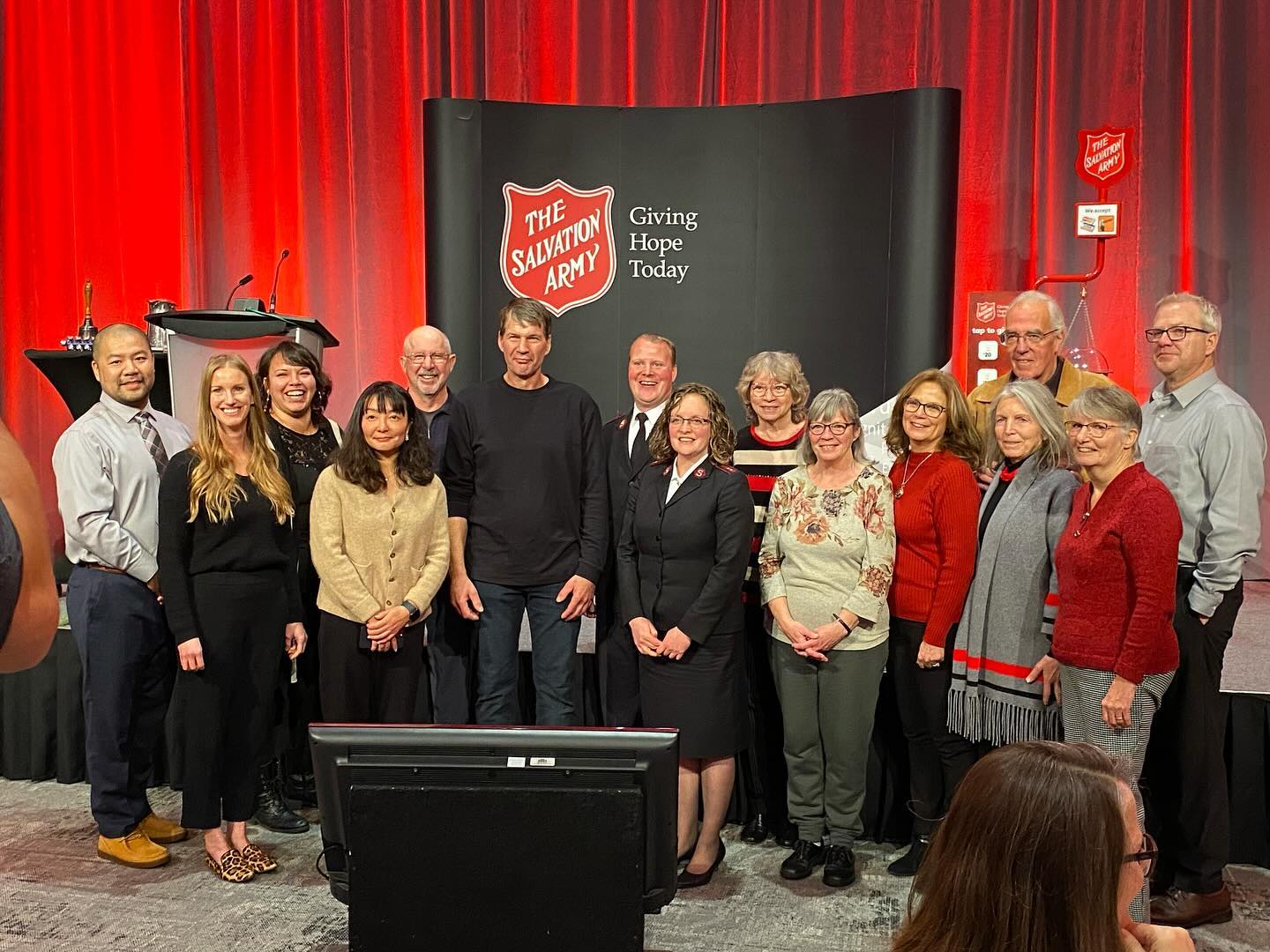 Group of people standing together on a stage in front of a Salvation Army backdrop and red curtain, posing for a group photo with podiums and presentation equipment visible nearby.