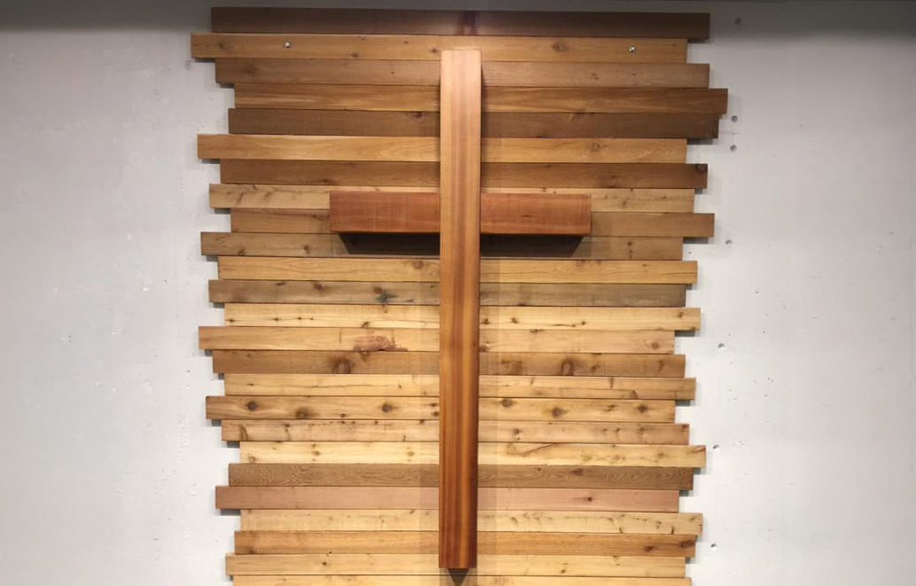 Wooden cross mounted on a wall made of horizontal wooden planks, centered against a light-colored background as part of a church interior display.